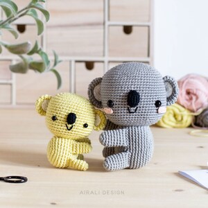 May include: Two crocheted koala plushies, one gray and one yellow, sitting on a wooden surface. The gray koala is larger than the yellow koala and both have black eyes and a smiling mouth.