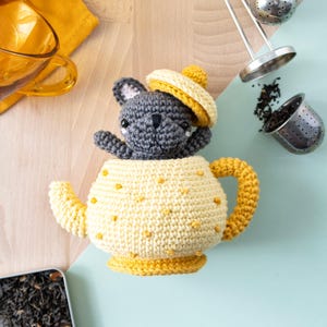 Könnte beinhalten: Eine gehäkelte Teekanne mit einer grauen Tierfigur, die ein gelbes Barett trägt. Die Teekanne ist gelb mit gelben Tupfen und einem gelben Griff. Zwei Tee-Eier und lose Teeblätter sind ebenfalls zu sehen.
