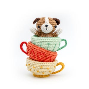 Può includere: Una pila di tre tazze da tè all'uncinetto in arancione, verde e giallo, sormontata da un cane all'uncinetto marrone e bianco. Le tazze da tè hanno dettagli decorativi e il cane sbuca dalla tazza superiore. Lo sfondo è bianco.