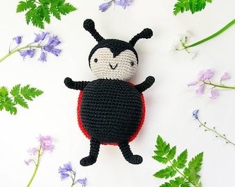 May include: A crocheted ladybug toy with black and red details, lying on a white background with small blue and white flowers.