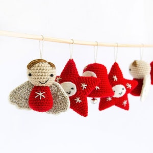 Pode incluir: Uma cole&ccedil;&atilde;o de enfeites de Natal feitos &agrave; m&atilde;o em croch&ecirc;. Os enfeites incluem um anjo sorridente, estrelas vermelhas com flocos de neve brancos e um sino vermelho. Est&atilde;o suspensos em uma vareta de madeira sobre um fundo branco.