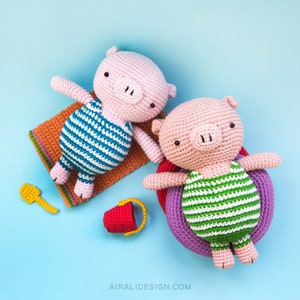 Puede incluir: Dos juguetes de cerdo de crochet que llevan trajes de ba&ntilde;o a rayas. Un cerdo est&aacute; acostado en una toalla de playa con una pala amarilla, mientras que el otro flota en un flotador inflable morado con un cubo rojo.