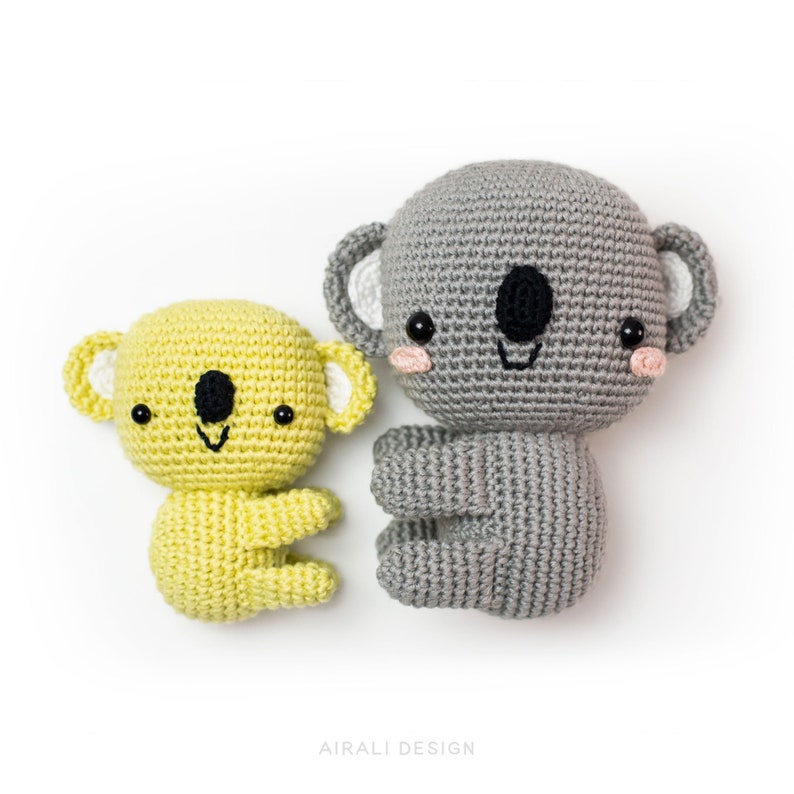 May include: Two crocheted koala plushies, one gray and one yellow, with black eyes and pink cheeks. The larger gray koala is sitting up, while the smaller yellow koala is lying down. The text "AIRALI DESIGN" is visible at the bottom of the image.