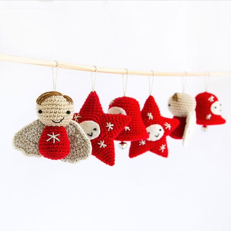 Pode incluir: Uma cole&ccedil;&atilde;o de enfeites de Natal feitos &agrave; m&atilde;o em croch&ecirc;. Os enfeites incluem um anjo sorridente, estrelas vermelhas com flocos de neve brancos e um sino vermelho. Est&atilde;o suspensos em uma vareta de madeira sobre um fundo branco.