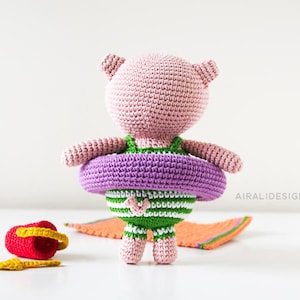 Puede incluir: Un cerdo de crochet rosa con un traje de ba&ntilde;o morado y verde y un flotador inflable morado. El cerdo est&aacute; de pie sobre una superficie blanca con un cubo rojo y una pala amarilla.
