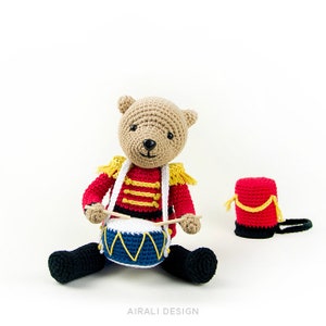 K&ouml;nnte beinhalten: Ein geh&auml;kelter Teddyb&auml;r, der als Schlagzeuger verkleidet ist, tr&auml;gt eine rot-goldene Uniform, h&auml;lt Schlagst&ouml;cke und sitzt vor einer blau-gelben Trommel. Eine rote geh&auml;kelte Trommel befindet sich rechts im Bild.