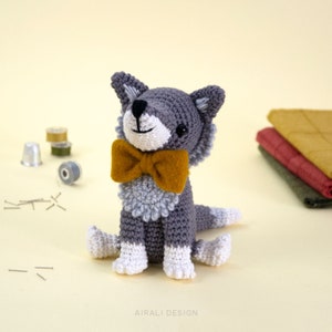 May include: A gray crocheted wolf wearing a yellow bow tie. The wolf is sitting with its paws tucked under its body. The wolf is a handmade amigurumi toy.