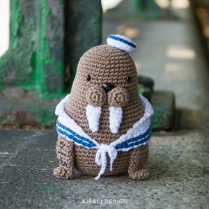 May include: A crocheted walrus wearing a sailor hat and a blue and white striped vest. The walrus has white tusks and brown fur.