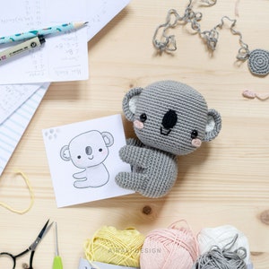 May include: A gray crocheted koala toy with a black nose and pink cheeks. The toy is sitting on a wooden surface with yarn, scissors, and a crochet hook.