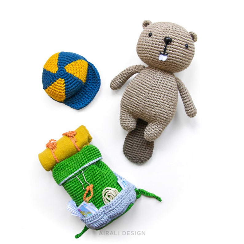 May include: A crocheted beaver wearing a blue and yellow hat, holding a green and yellow backpack. The backpack is filled with a yellow sleeping bag, a green rope, and a blue map.
