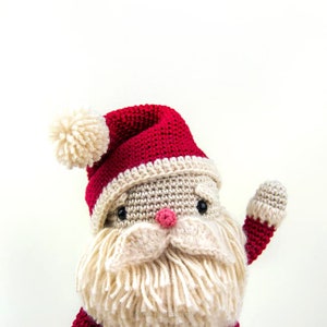 May include: A crocheted Santa Claus doll with a red hat and white pom-pom, a white beard, and black eyes. The doll is waving with its right arm.
