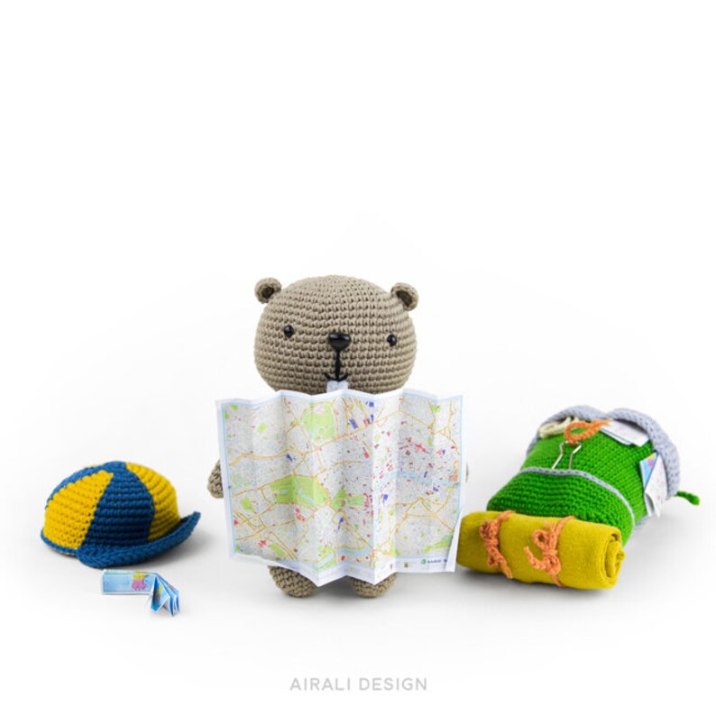 May include: A crocheted brown bear holding a map, wearing a blue and yellow hat, with a green sleeping bag and a yellow sleeping mat on a white background.  A small blue and yellow map is on the ground.