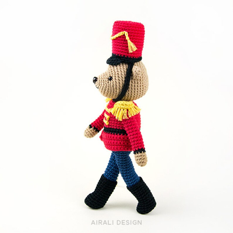 K&ouml;nnte beinhalten: Ein geh&auml;keltes Spielzeugb&auml;r, der als Spielzeugsoldat verkleidet ist. Der B&auml;r tr&auml;gt eine rot-schwarze Uniform mit goldenen Verzierungen, einen roten Hut mit goldener Quaste und schwarze Stiefel.