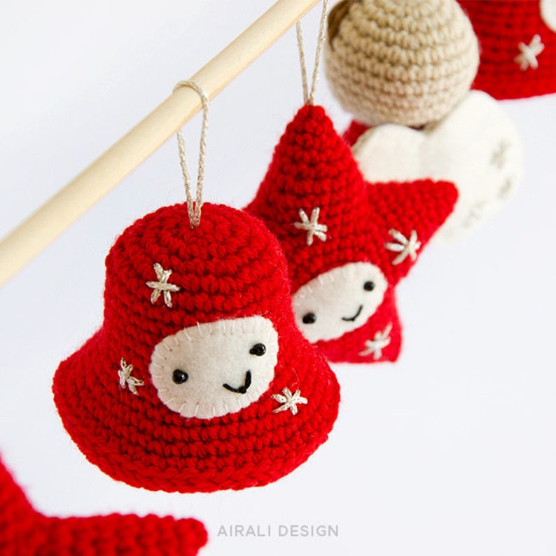 Pode incluir: Close-up de enfeites de Natal de croch&ecirc; vermelho feitos &agrave; m&atilde;o, incluindo uma forma de sino e estrela, com rostos de feltro branco e flocos de neve bordados. Os enfeites est&atilde;o pendurados em um pino de madeira. O tema &eacute; decora&ccedil;&atilde;o de f&eacute;rias.