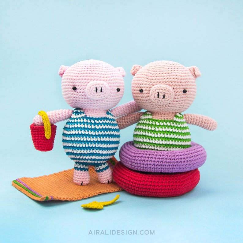 Puede incluir: Dos juguetes de cerdo de crochet que llevan trajes de ba&ntilde;o. Un cerdo sostiene un cubo rojo y est&aacute; de pie sobre una toalla de playa naranja a rayas. El otro cerdo est&aacute; sentado en un flotador inflable morado y rojo.
