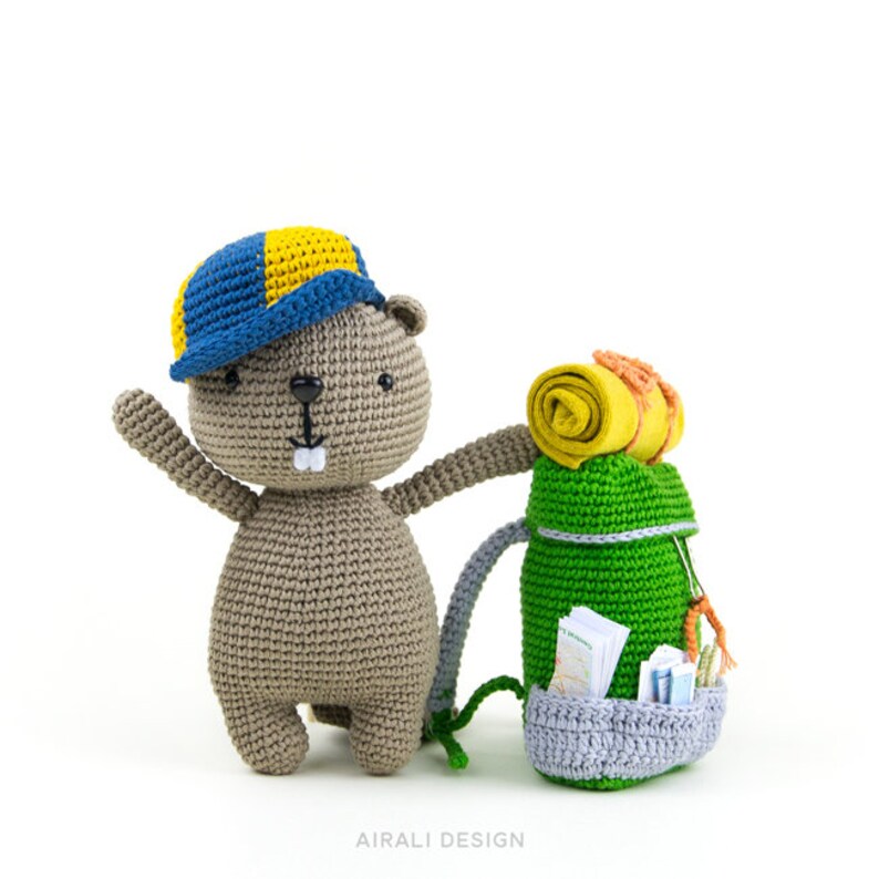 May include: A crocheted gray beaver wearing a blue and yellow hat, holding a green crocheted backpack with a rolled-up orange sleeping bag on top. The backpack has a gray bottom and contains a few small crocheted items.