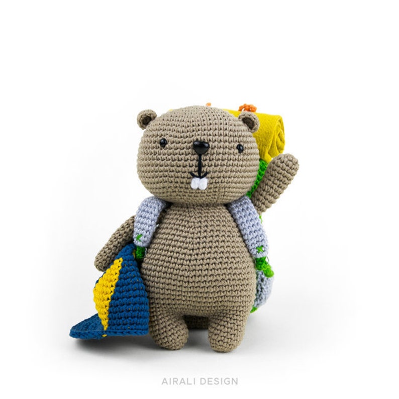 May include: A crocheted brown bear wearing a blue and yellow hat and a green and blue backpack. The bear is smiling and has a white tooth showing. The bear is standing on a white background.
