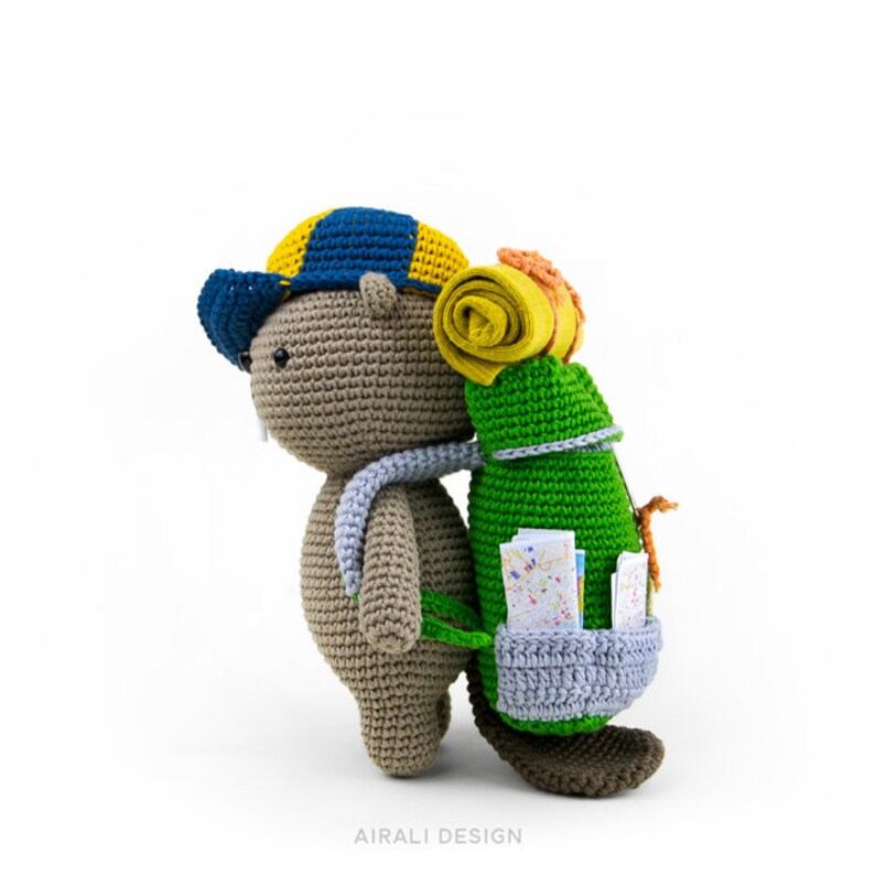 May include: A crocheted stuffed animal beaver wearing a blue and yellow hat and carrying a green backpack with a rolled-up yellow sleeping bag and a map.