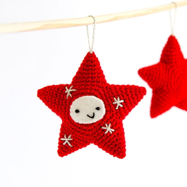 Pode incluir: Enfeite de estrela vermelha de croch&ecirc; com um rosto sorridente e detalhes bordados. A estrela tem um rosto de feltro branco e pequenos detalhes de estrelas brancas. Uma corda prateada &eacute; presa para pendurar. Uma segunda estrela vermelha &eacute; vis&iacute;vel.