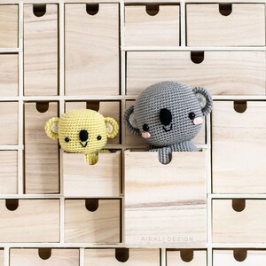May include: Two crocheted koala bears, one gray and one yellow, peeking out of wooden drawers. The drawers are part of a larger wooden storage unit.