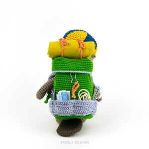 May include: A crocheted stuffed animal wearing a blue and yellow hat and a green backpack. The backpack is filled with crocheted items, including a sleeping bag, rope, and maps.
