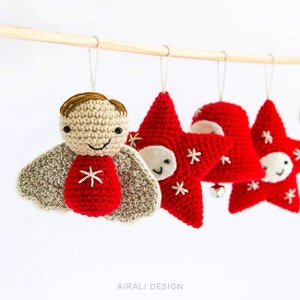Pode incluir: Enfeites de Natal de croch&ecirc; pendurados em uma vara de madeira. Os enfeites incluem um anjo sorridente com pele bronzeada, cabelo castanho e detalhes vermelhos, e enfeites vermelhos em forma de estrela com rostos sorridentes e flocos de neve brancos.