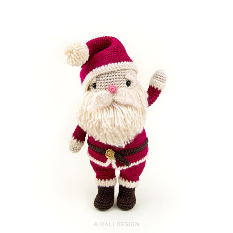 May include: A crocheted Santa Claus doll with a red hat, white beard, and brown boots. He is wearing a red and white suit with a brown belt and a gold buckle. He is waving with his right hand.