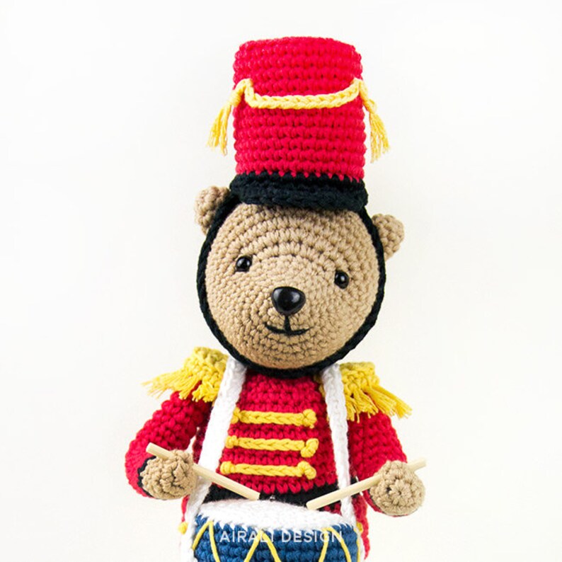 K&ouml;nnte beinhalten: Ein geh&auml;kelter Teddyb&auml;r, der als Schlagzeuger verkleidet ist, tr&auml;gt einen roten und schwarzen Hut und eine rote und gelbe Uniform. Der B&auml;r h&auml;lt Schlagst&ouml;cke und eine blaue und gelbe Trommel mit dem Text "AIRALI DESIGN" darauf.