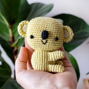 May include: A crocheted koala toy with a yellow body, white ears, and black eyes. The koala is sitting with its arms crossed.