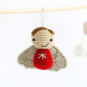 Pode incluir: Enfeite de anjo de croch&ecirc; com cabe&ccedil;a bege, cabelo castanho e corpo vermelho com uma estrela branca. O anjo tem asas cinzentas e um rosto sorridente. Ele pende de um pino de madeira com um fio prateado.