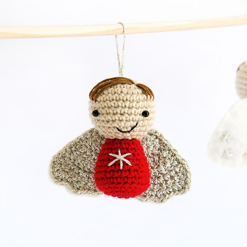 Pode incluir: Enfeite de anjo de croch&ecirc; com cabe&ccedil;a bege, cabelo castanho e corpo vermelho com uma estrela branca. O anjo tem asas cinzentas e um rosto sorridente. Ele pende de um pino de madeira com um fio prateado.