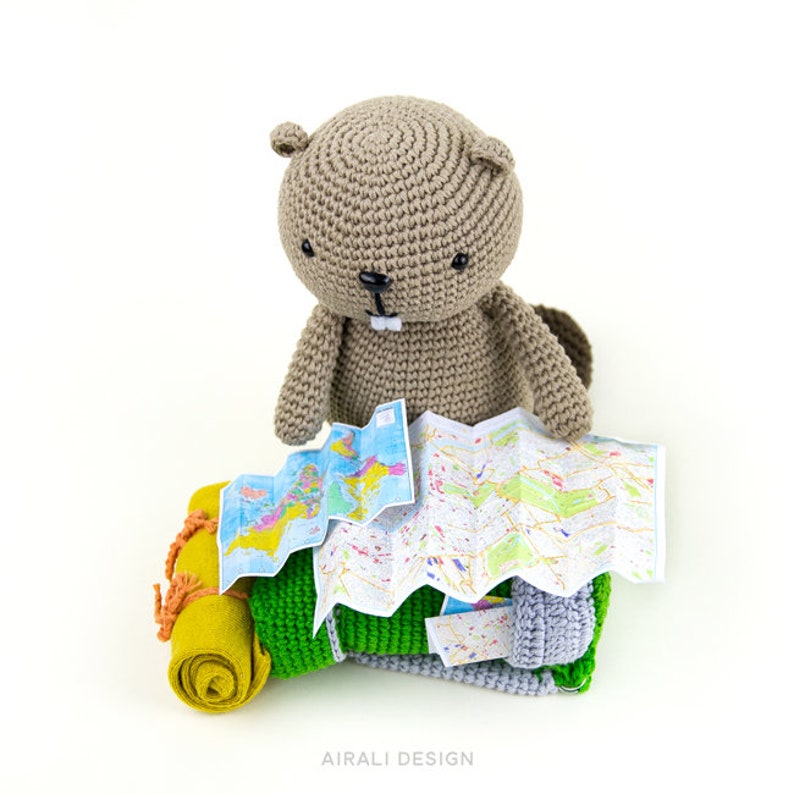 May include: A brown crocheted beaver plush toy sits on a green and yellow crocheted sleeping bag with a folded map on top.