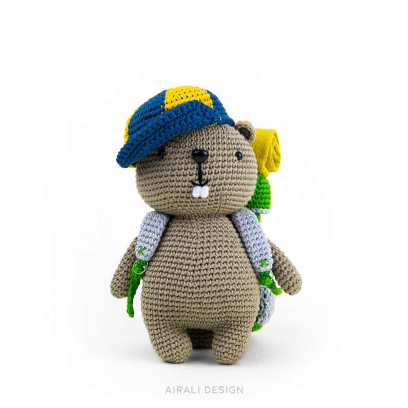 May include: A brown crocheted beaver wearing a blue and yellow hat and a green and yellow backpack. The beaver has a small, white smile.