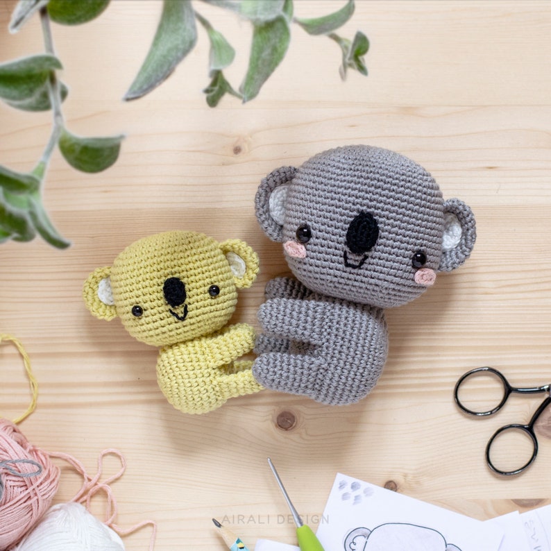 May include: Two crocheted koala plushies, one gray and one yellow, are sitting on a wooden surface. The gray koala is larger than the yellow koala. Both koalas have black eyes and a black nose. The gray koala has pink cheeks.