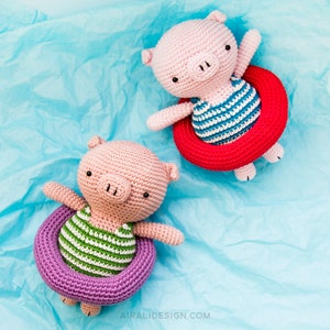 Puede incluir: Dos juguetes de cerdo de crochet que llevan trajes de ba&ntilde;o a rayas y flotan en flotadores inflables de colores. El cerdo de la izquierda lleva un traje de ba&ntilde;o a rayas verdes y blancas y un flotador morado. El cerdo de la derecha lleva un traje de ba&ntilde;o a rayas azules y blancas y un flotador rojo.