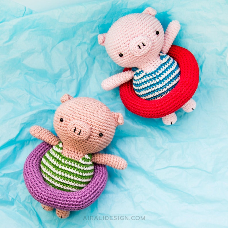 Puede incluir: Dos juguetes de cerdo de crochet que llevan trajes de ba&ntilde;o a rayas y flotan en flotadores inflables de colores. El cerdo de la izquierda lleva un traje de ba&ntilde;o a rayas verdes y blancas y un flotador morado. El cerdo de la derecha lleva un traje de ba&ntilde;o a rayas azules y blancas y un flotador rojo.