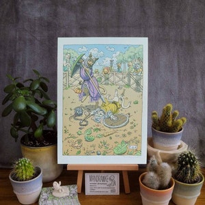 May include: A colourful illustration of a cat in armour wielding a spear, fighting another cat in a shield and armour. The illustration is printed on white paper and displayed on a wooden easel. The image also includes a business card with the text "MANDRAWKE" and a small white ceramic cat figurine.
