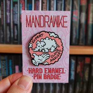 May include: A pink and white hard enamel pin badge with the text "MANDRAWKE" at the top. The pin features a cloud design with the words "EXPERT CLOUD ANALYST". The bottom text reads "HARD ENAMEL PIN BADGE".