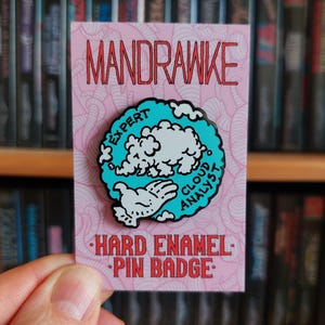 May include: A pink hard enamel pin badge with the word "MANDRAWKE" in red. The pin features a blue and white cloud design with the text "EXPERT CLOUD ANALYST". The words "HARD ENAMEL PIN BADGE" are also in red.
