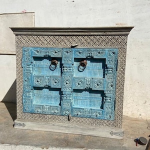 May include: A distressed white wooden cabinet with two doors. The doors are painted a light blue and have intricate carved details. The cabinet has two metal ring handles.