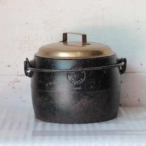以下が含まれることがあります： 真鍮の蓋とハンドルが付いた、黒い鋳鉄製のヴィンテージポット。 ポットは丸い形で、両側に2つのハンドルと前面に装飾的なエンブレムが付いています。 蓋には真鍮製のハンドルが付いています。