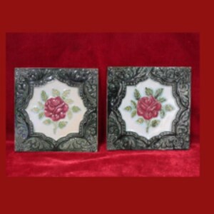 May include: Two square decorative tiles with a floral design. Each tile features a red rose in the center, surrounded by green leaves, set against a white background. The tiles are framed with a dark, ornate border.