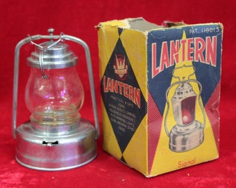 Può includere: Una lanterna vintage in argento con un globo di vetro e un manico. La lanterna è nella sua scatola originale, che è gialla, rossa e blu con la parola "LANTERN" stampata in grandi caratteri grassi. La scatola ha anche il testo "Signal" stampato sul fondo.
