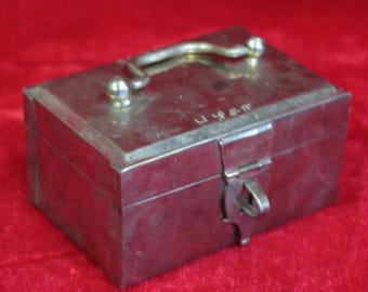 Pode incluir: Uma pequena caixa retangular de metal prateado com tampa articulada e alça. A caixa tem um fecho e pequenas esferas decorativas na alça e na tampa. Em um fundo vermelho.