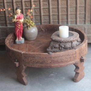 May include: A round wooden table with intricate carvings and three legs. The table has a raised edge and a decorative design around the rim. A small statue of a woman in a red dress and a white candle on a wooden base are on the table.