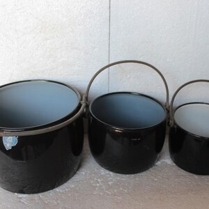 Könnte beinhalten: Drei schwarze Emaille-Töpfe unterschiedlicher Größe mit Metallgriffen. Die Töpfe haben einen grauen Innenraum. Diese Töpfe sind wahrscheinlich zum Kochen oder für dekorative Zwecke gedacht.