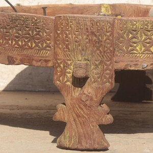 Pu&ograve; includere: Un tavolo rotondo in legno con intagli intricati e una macchia gialla. Il tavolo ha tre gambe intagliate in uno stile decorativo.