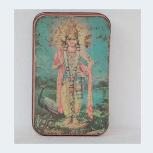Pode incluir: Caixa de lata retangular vintage com uma ilustração colorida de uma divindade e um pavão. A obra de arte apresenta uma figura em roupas ornamentadas, uma coroa e uma grinalda, sobre um fundo turquesa. A lata tem uma aparência envelhecida e desgastada.