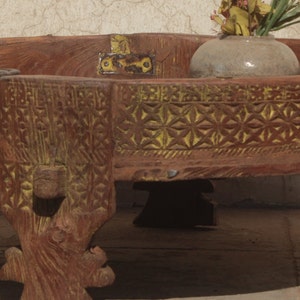 Pu&ograve; includere: Un tavolo rotondo in legno con intagli intricati e una finitura giallo-marrone. Il tavolo ha tre gambe e una cerniera in metallo decorativa sulla parte superiore.