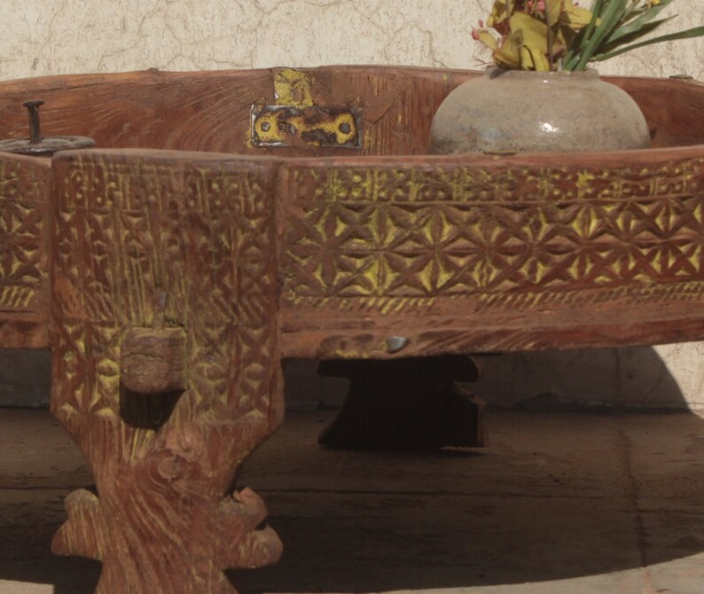 Pu&ograve; includere: Un tavolo rotondo in legno con intagli intricati e una finitura giallo-marrone. Il tavolo ha tre gambe e una cerniera in metallo decorativa sulla parte superiore.